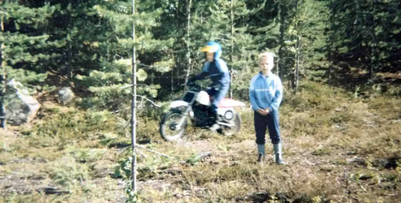 Two kids in a forest with one of them riding on a dirt bike