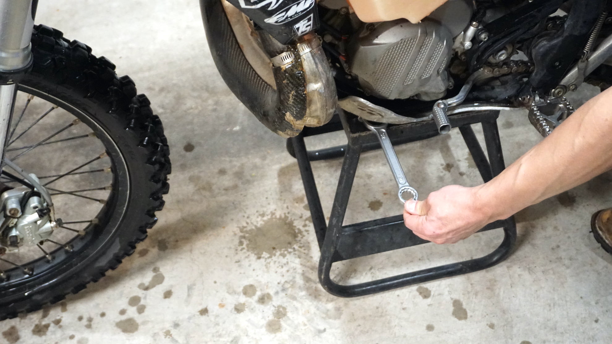 Dirt bike on a center stand with hand pushing a wrench under the skid plate
