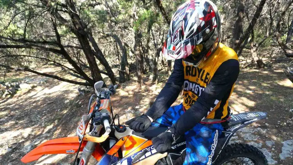 A rider in full riding gear showing forearms in arm pump with fists closed while sitting on a dirt bike on a forest trail