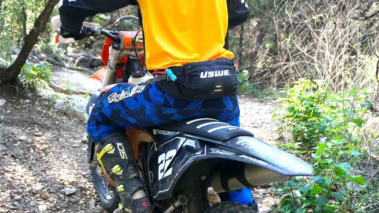 A man sitting on a dirt bike facing away with left leg on the foot peg and sitting down on a ribbed seat
