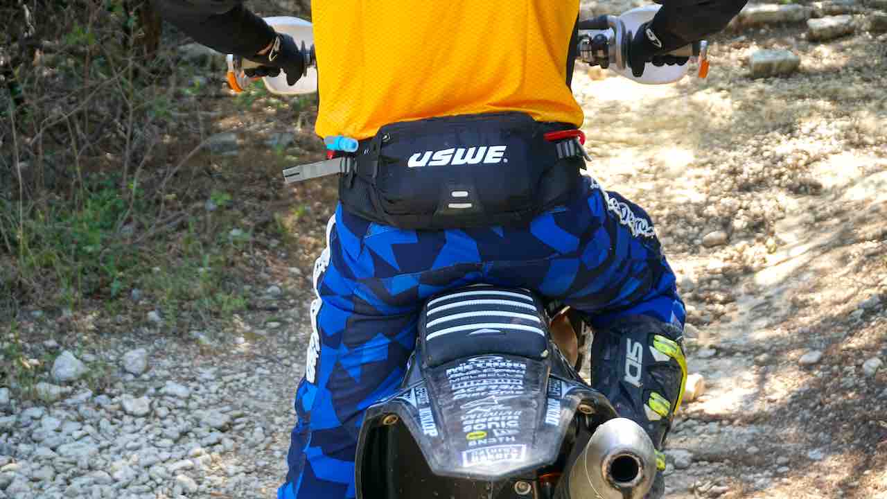 A dirt bike rear view with a man sitting on a black and white seat and wearing a waist hydration pack