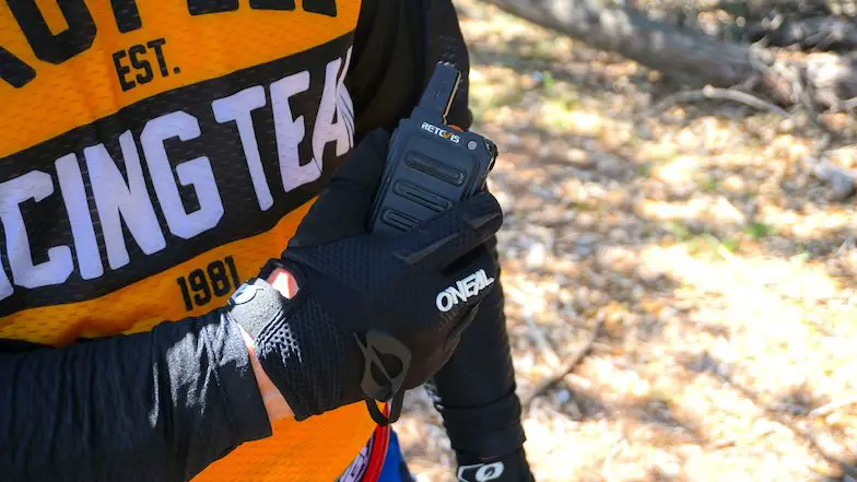 Rider with a walkie talkie in right hand with gloves on and a yellow dirt bike jersey on the background