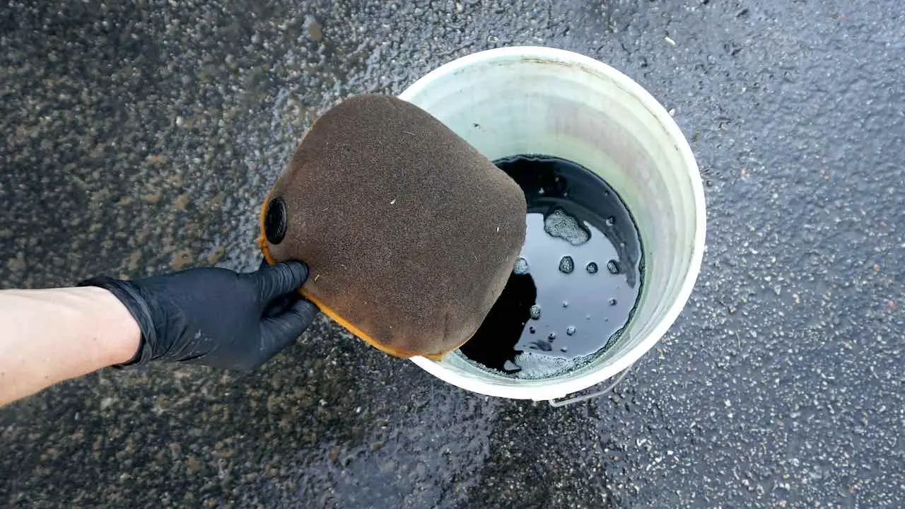 A hand holding a dirty dirt bike air filter above a bucket filled with a air filter cleaner