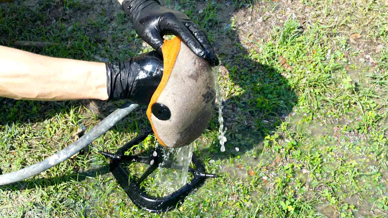Hands holding a dirt bike air filter and rinsing it with a water hose while the air filter cage is laying on the ground
