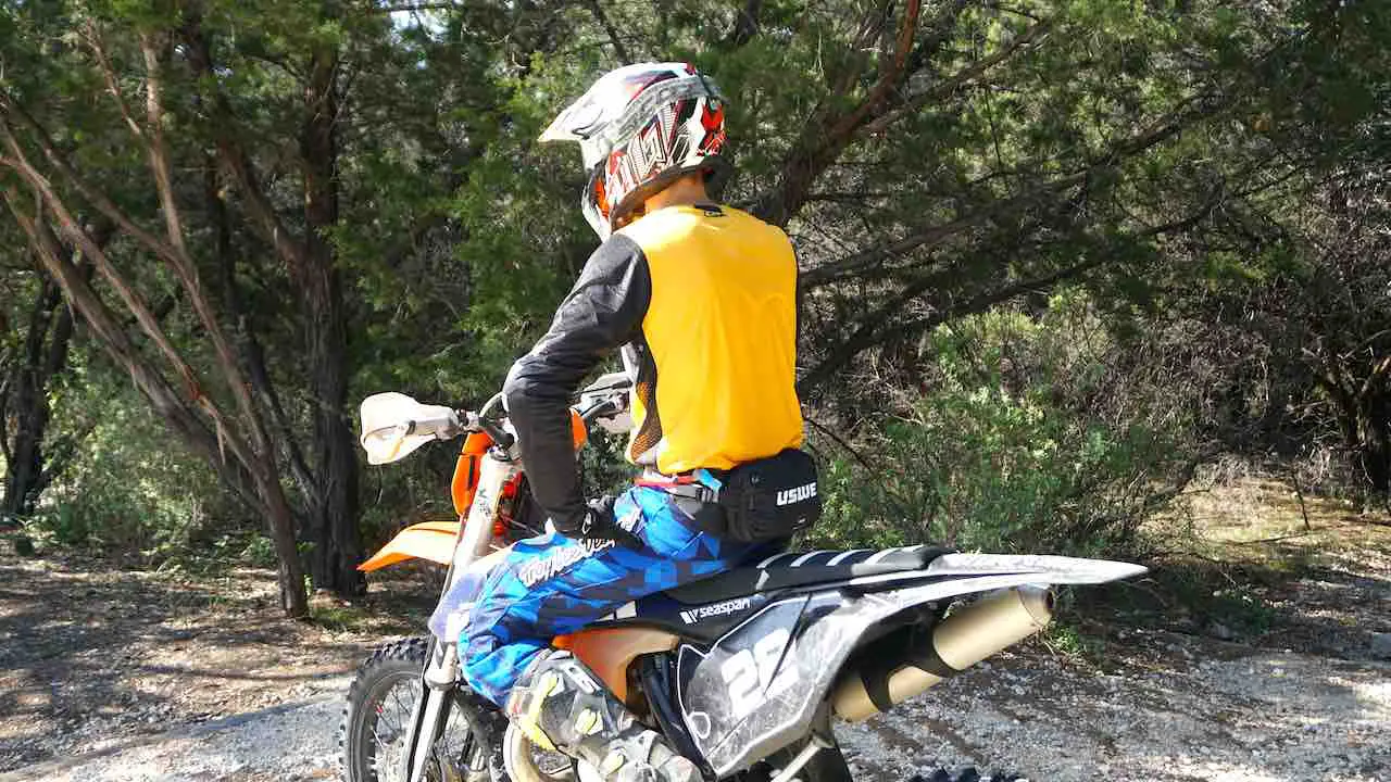 A man sitting on a dirt bike facing away with yellow jersey, blue pants and fanny pack on