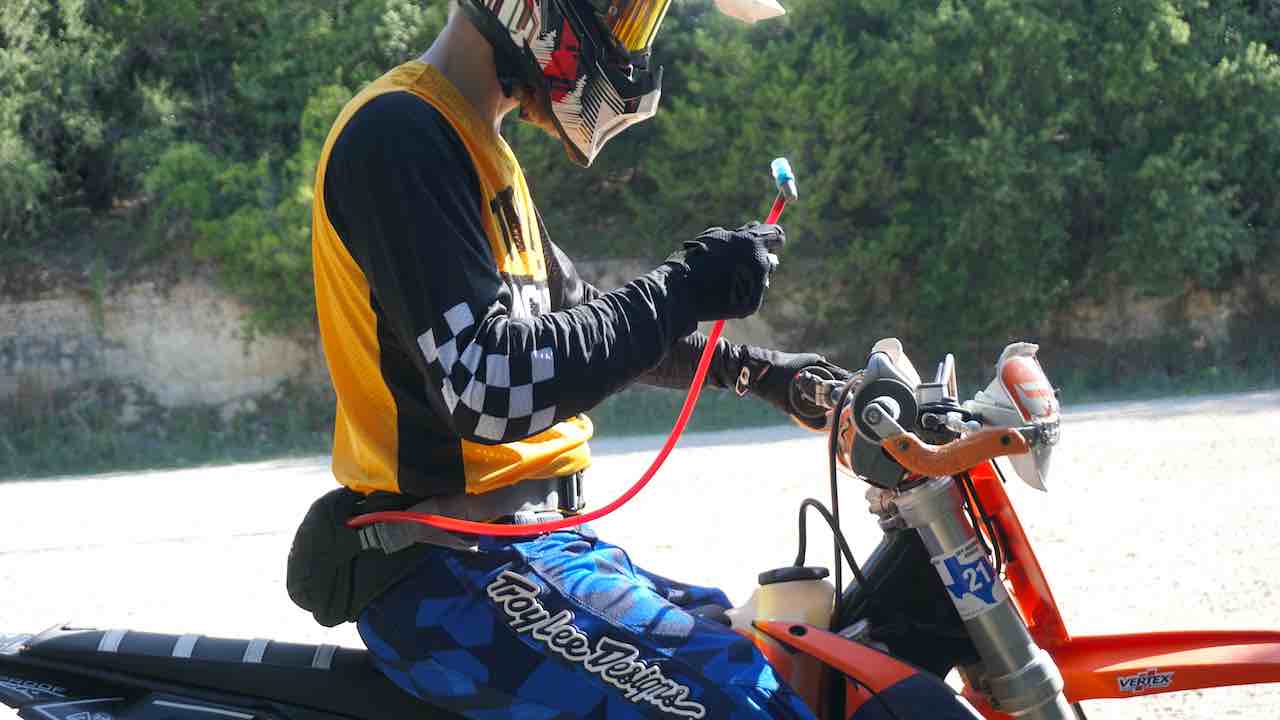 A dirt bike rider sitting on a dirt bike wearing a hydration fanny pack and holding a drinking tube in one hand