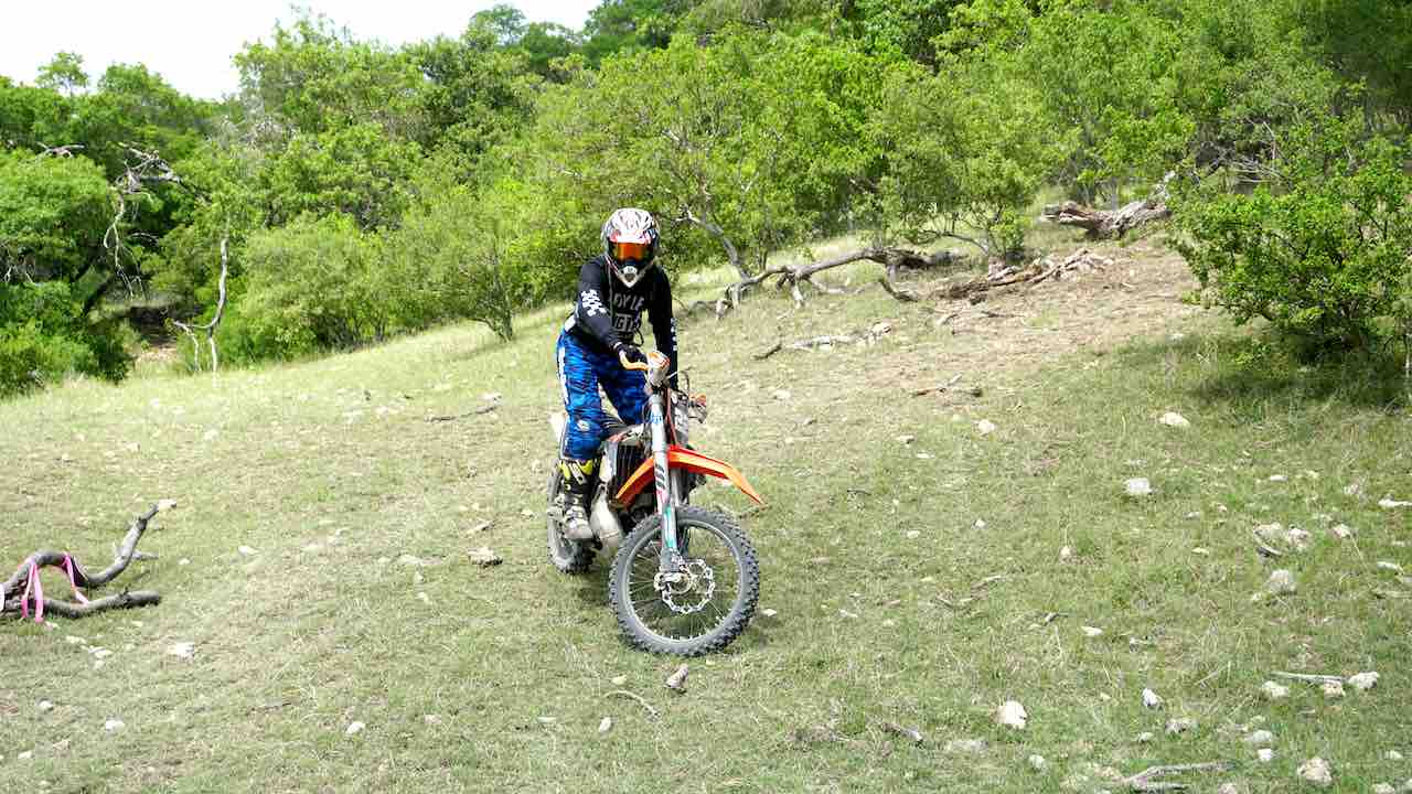 A man wearing full riding gear and balancing on a dirt bike on a grassy hill