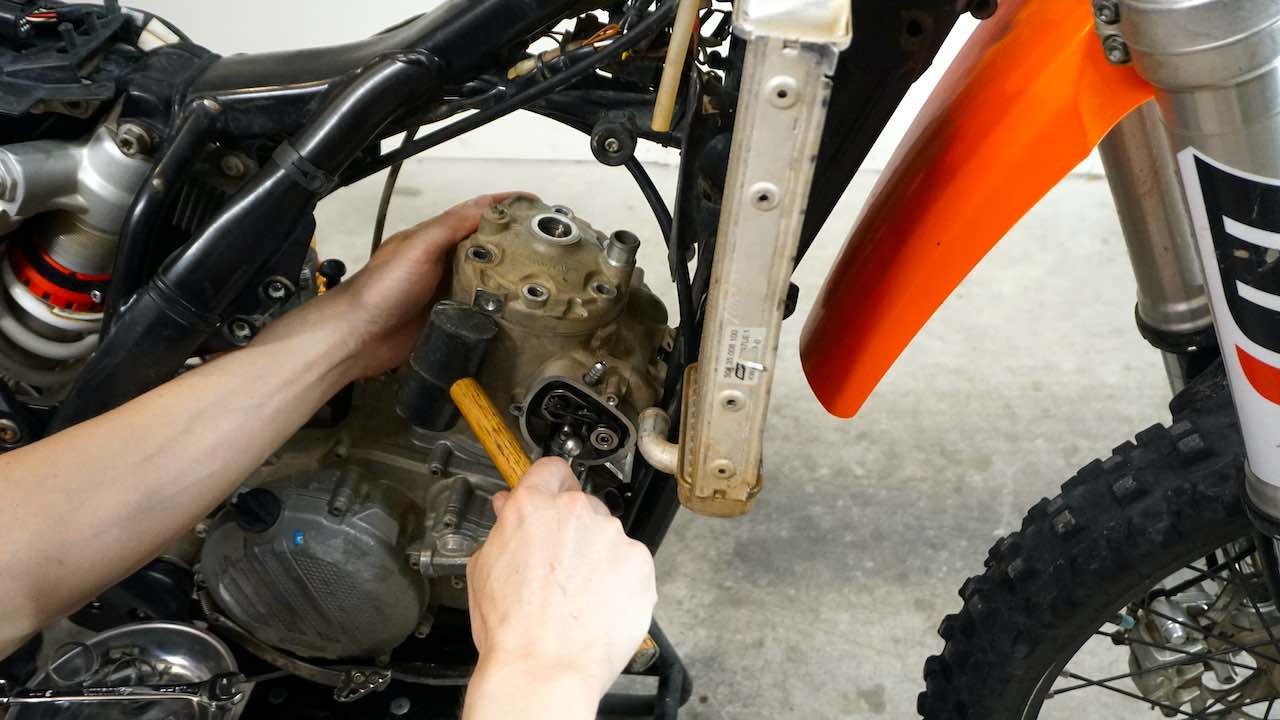 A hand holding the dirt bike cylinder head and tapping it with a rubber hammer