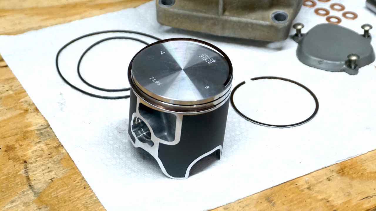 A bottom piston ring almost installed in the piston slot with other 2 stroke top end rebuild parts laying on a paper towel