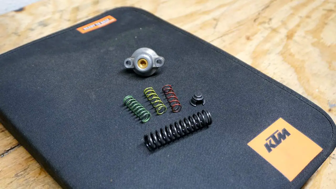 Power valve cover, green, yellow and red auxiliary springs and an adjusting spring laying on top of a KTM owner's manual bag