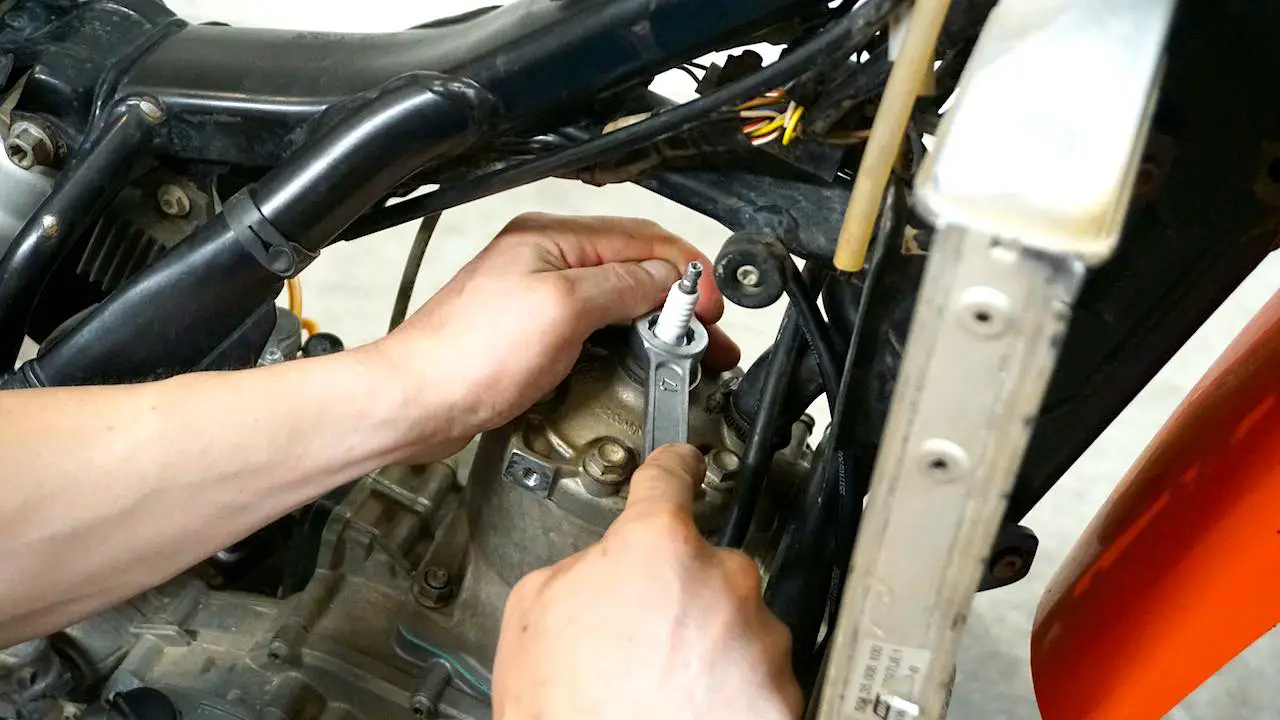 Hands holding a wrench around a white spark plug on a dirt bike cylinder head and about to change the spark plug