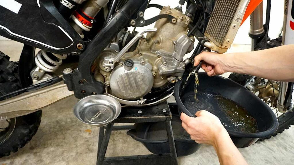 Hands holding a drain pan and draining coolant from a radiator hose on the right side of a dirt bike under the right radiator