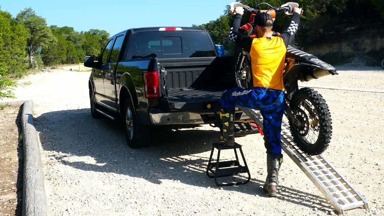 A man holding a dirt bike by the handlebars and pushing it up a ramp into a pickup truck with left foot on a center stand