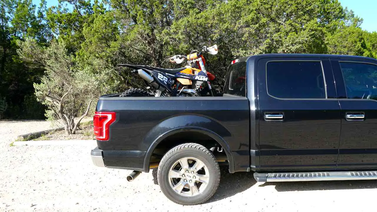 A dirt bike loaded onto a black pickup truck bed and fully strapped down with a forest on the background
