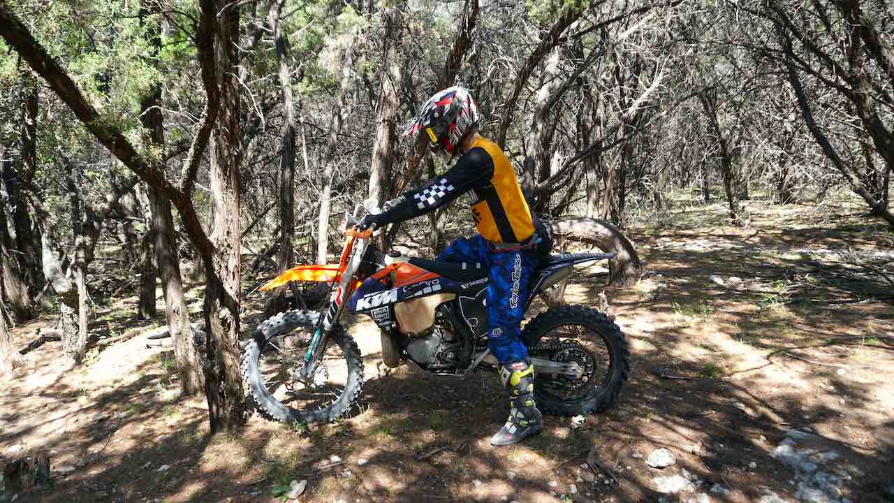 Rider getting ready to stand on a dirt bike with front tire against a tree